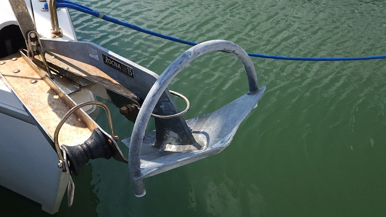 Close-up of a Rocna anchor mounted on the bow roller of a sailboat, with green water in the background