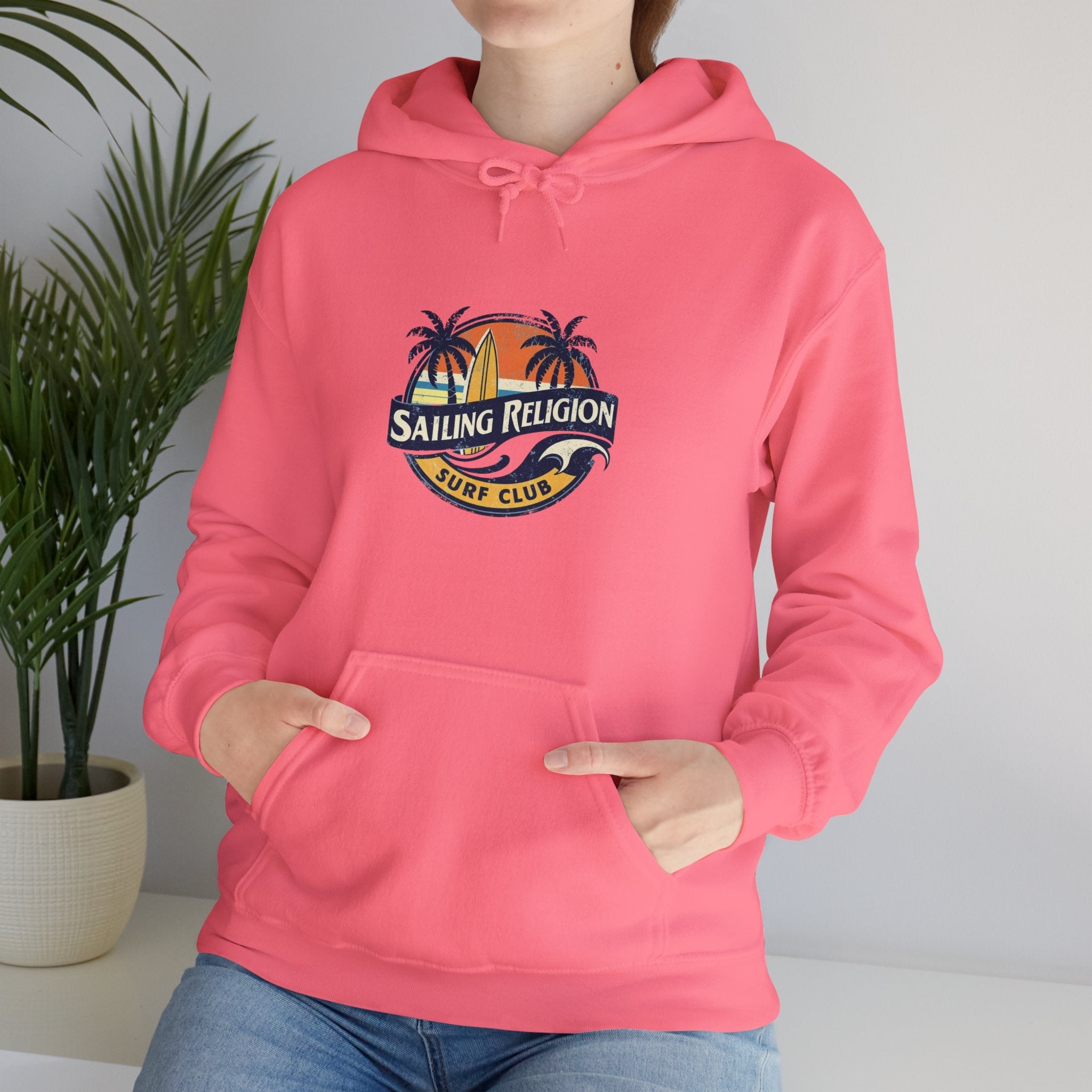 Woman wearing a pink Sailing Religion hoodie featuring a retro surf club logo with palm trees, sunset, and surfboard — cozy ocean-inspired pullover for beach lovers and coastal lifestyle.